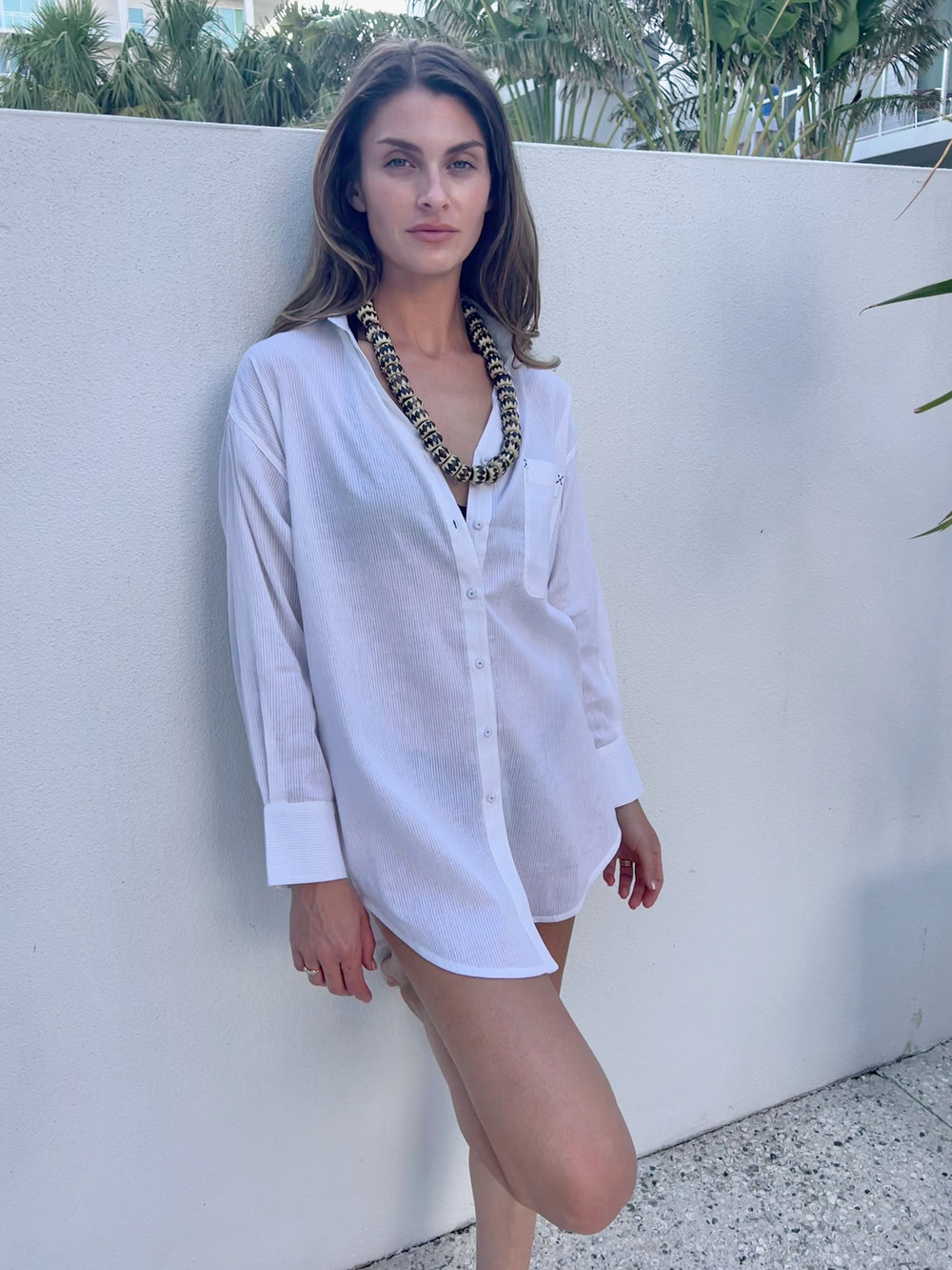 A woman wearing a white oversized shirt with a subtle stripe pattern, featuring stitched-down pintucks and embroidery in a neutral setting.
