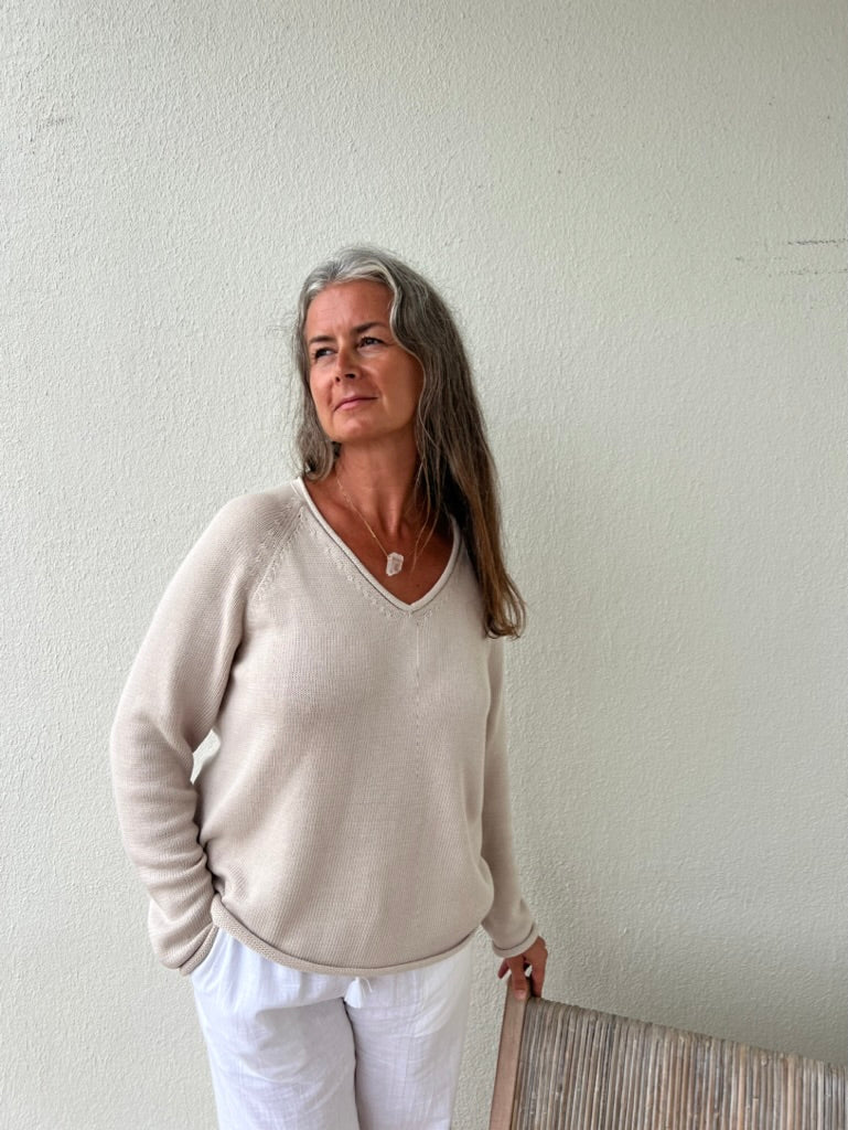 Woman wearing a beige sweater and white pants against a plain wall.