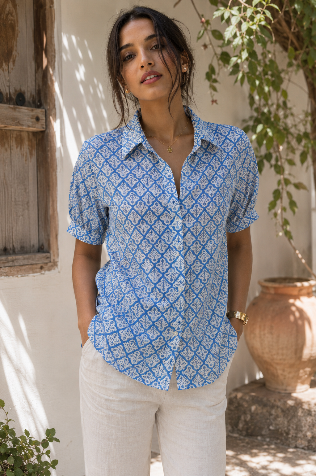 Woman wearing a blue patterned shirt standing outdoors with a white wall and potted plant in the background.