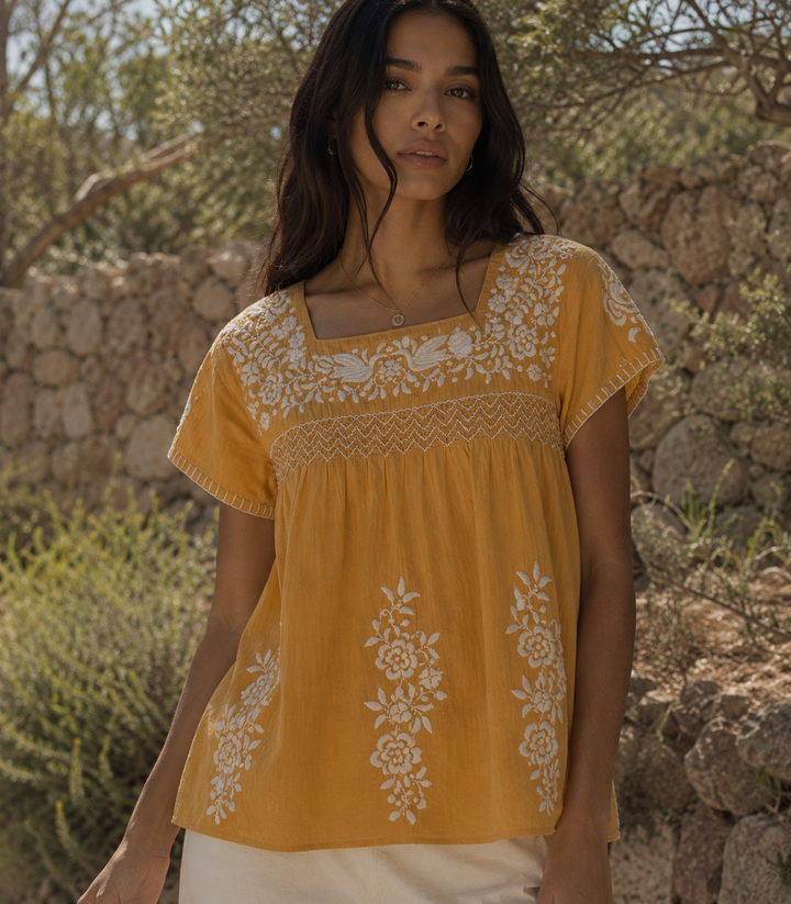 Woman wearing a yellow embroidered top standing in front of a stone wall and greenery.
