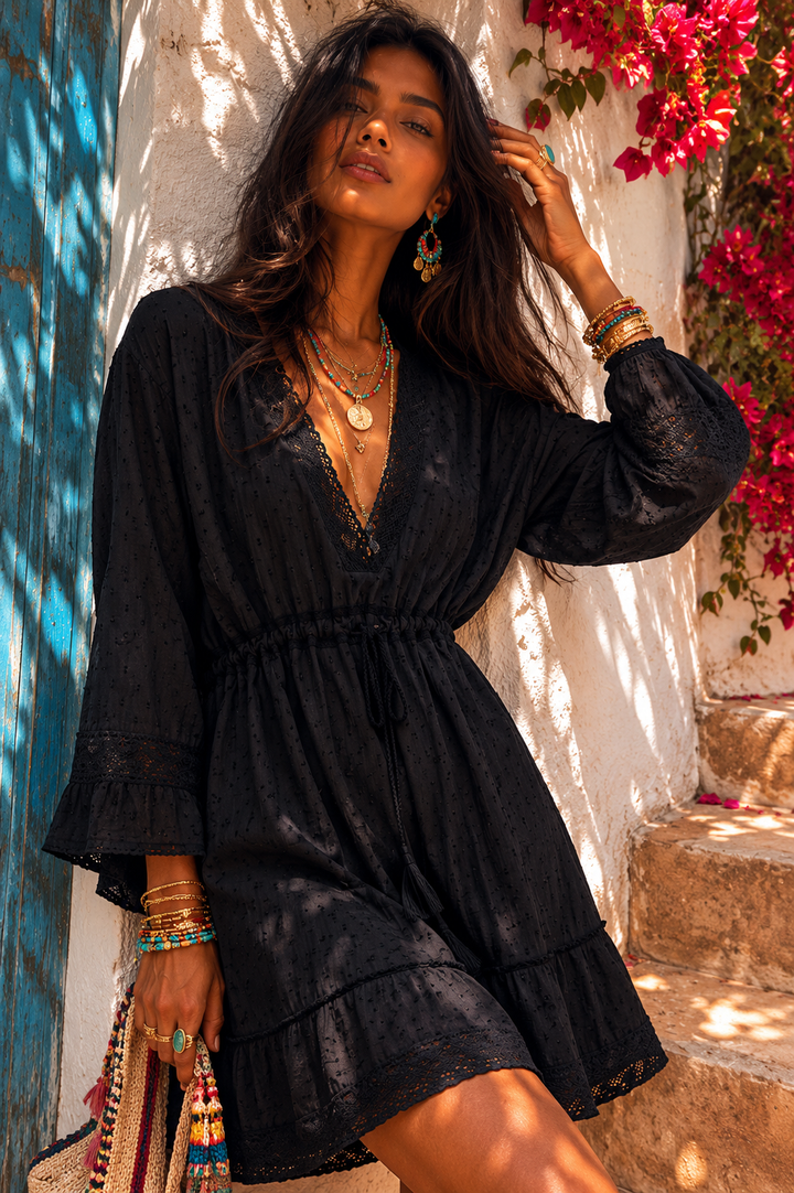 Woman in a black dress standing in a sunlit outdoor setting with flowers and a blue wall.
