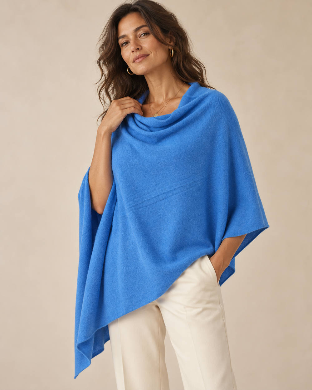 A woman wearing a bright blue knit poncho draped over her shoulders, paired with off-white trousers, against a plain beige studio background.