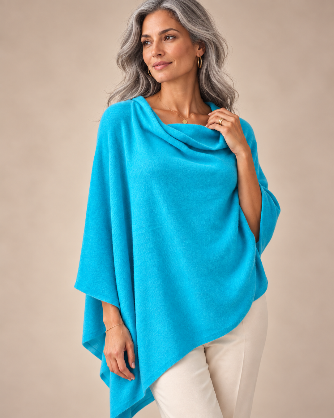 A woman with gray hair models a bright turquoise blue poncho-style wrap over cream pants, set against a solid beige studio background.