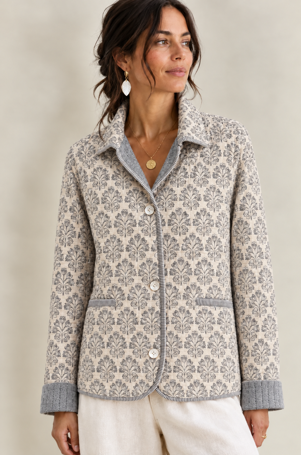 Model wearing an ivory floral quilted jacket with soft gray contrast lining and cuffs, styled with white pants and gold jewelry against a neutral studio background.