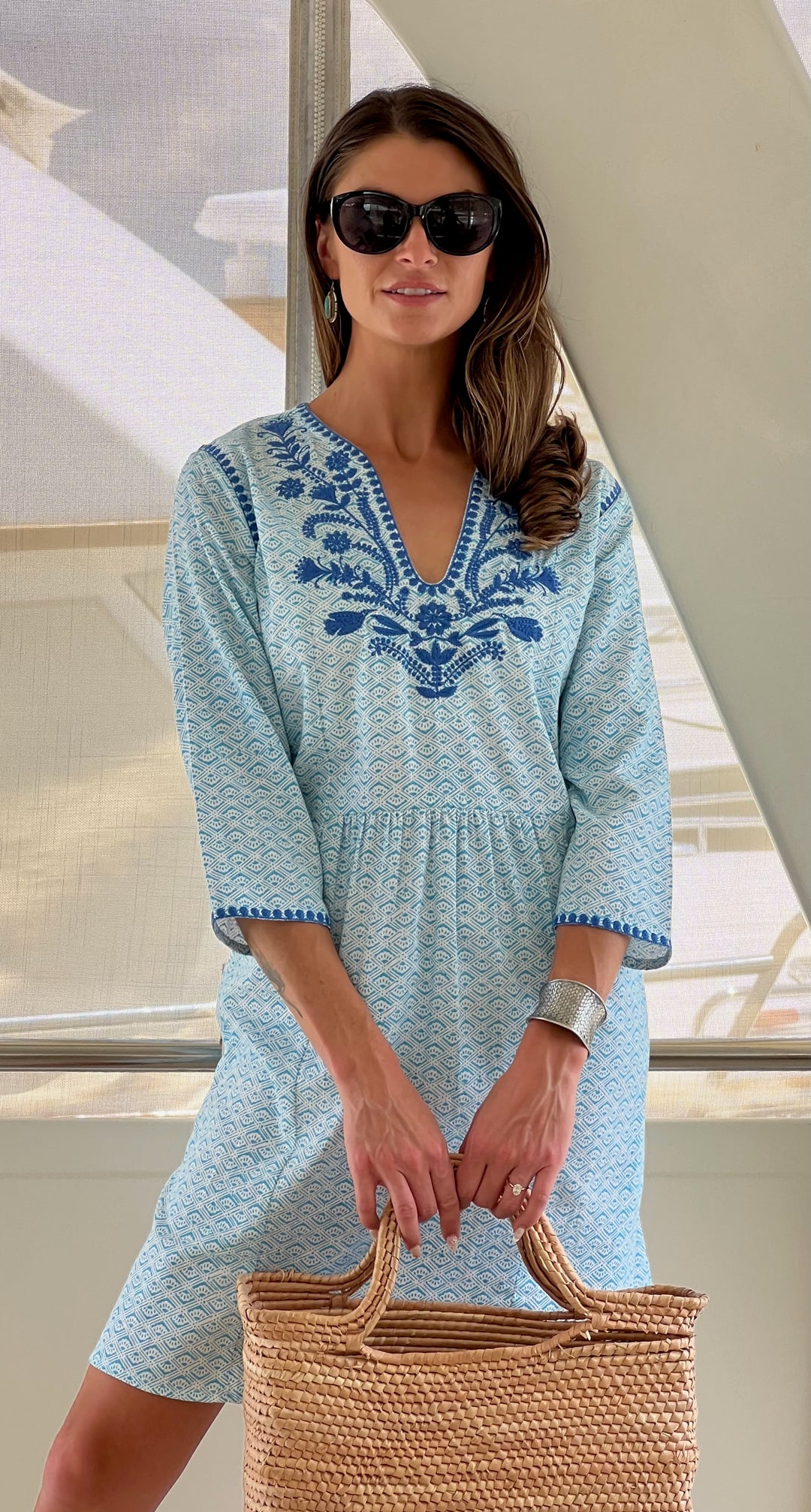 A woman wearing a knee-length blue cotton dress with white trellis print blockprint and bird & flower embroidery, sunglasses, and carrying a straw tote bag.
