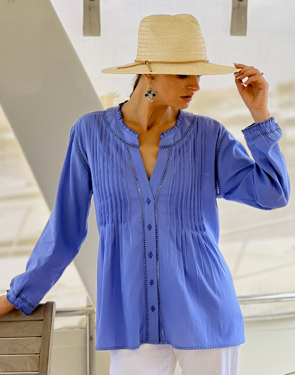 A woman wearing a blue long-sleeve cotton blouse with lace details and pin-tucked patterns, paired with white pants, accessorized with earrings and a wide-brimmed hat.