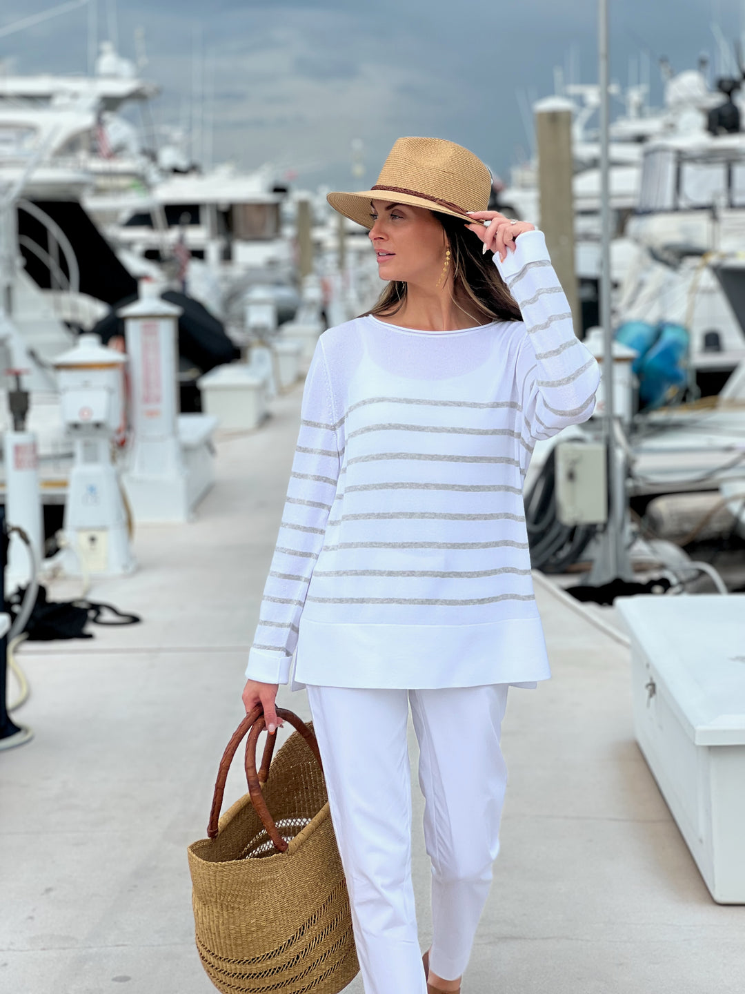 Woman in a striped sweater and white pants standing on a dock with boats in the background