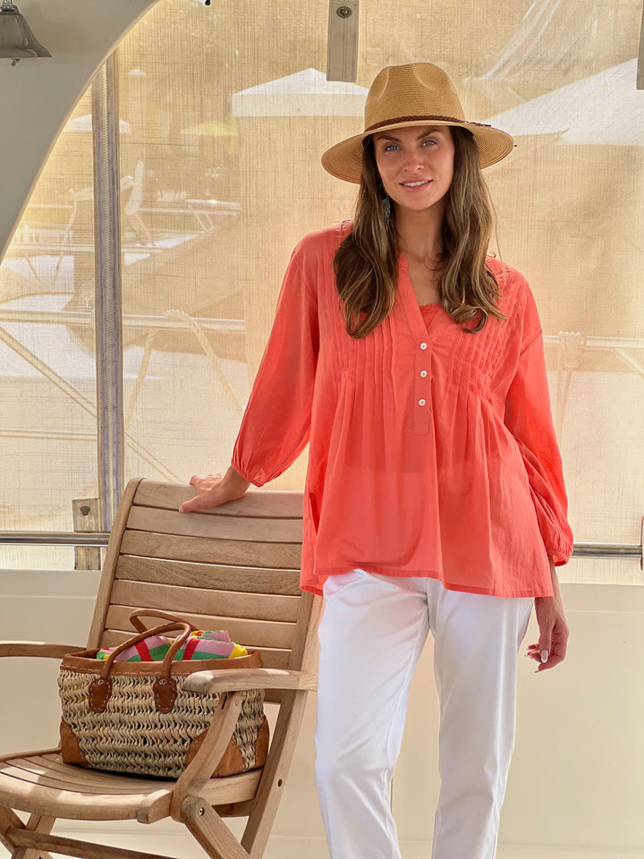 Woman in a coral shirt and white pants standing next to a wooden chair with a straw bag on a deck.