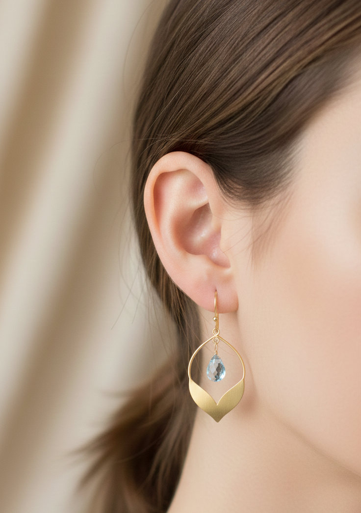 Gold earrings with a blue gemstone worn by a person against a beige background