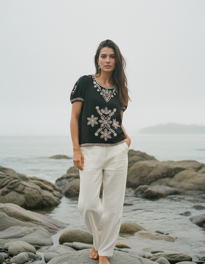 Woman standing on a rocky beach wearing a black embroidered top and white pants.