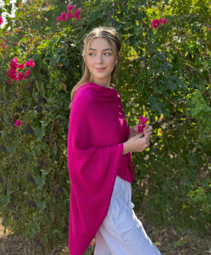 Woman in a bright pink shawl standing in front of green foliage with pink flowers.
