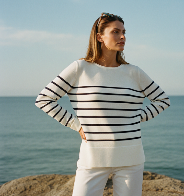 Woman wearing a black and white striped sweater and white pants standing on a rocky outcrop with ocean and sky in the background.