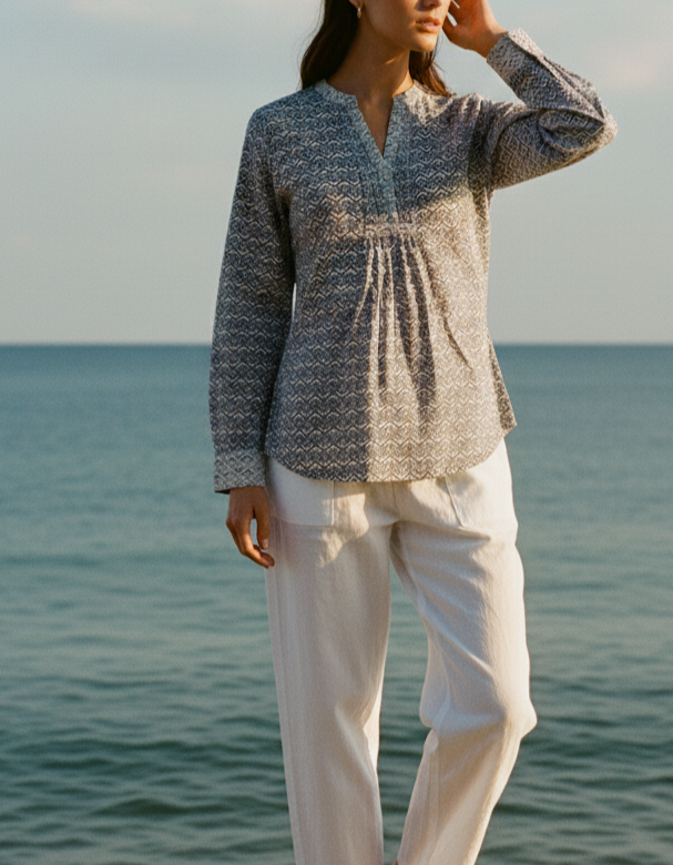 Woman standing on a rock by the ocean wearing a patterned shirt and white pants.
