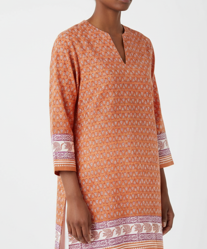 Person wearing an orange patterned tunic with white pants on a light gray background