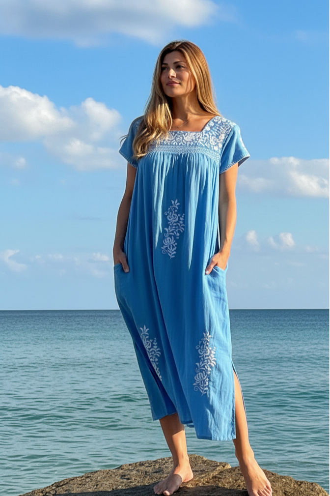 Woman in a blue cotton dress standing on a rock by the ocean with a clear sky.