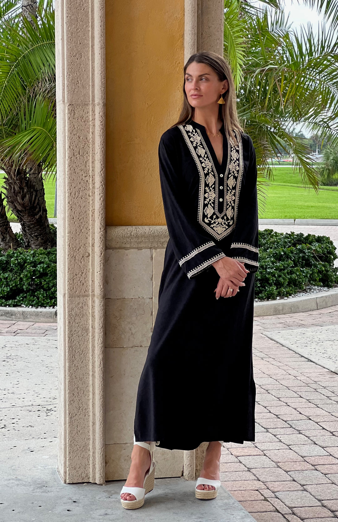 Woman in a black dress with white embroidery standing outdoors near a building.