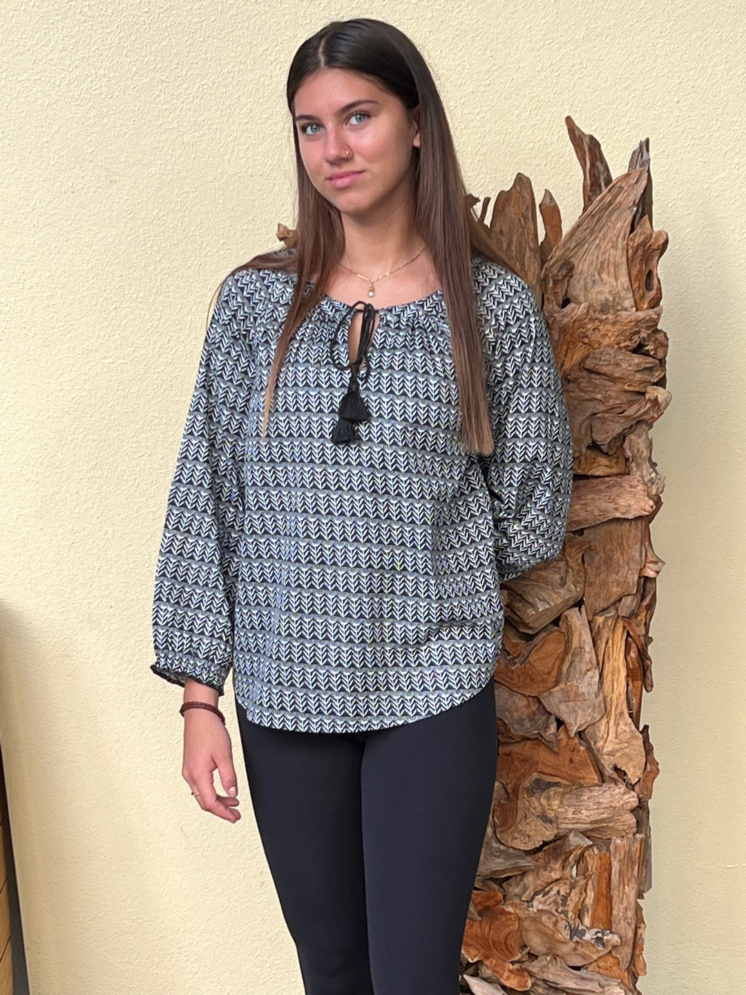 Woman wearing a patterned blouse standing next to a wooden log against a beige wall.