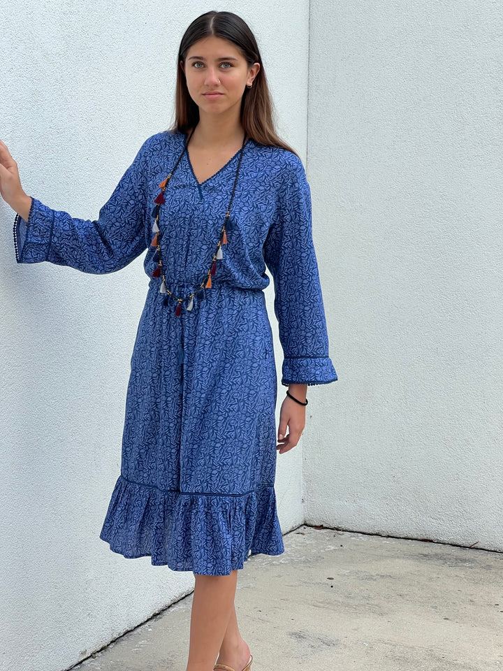 Woman wearing a blue dress with long sleeves against a white wall.