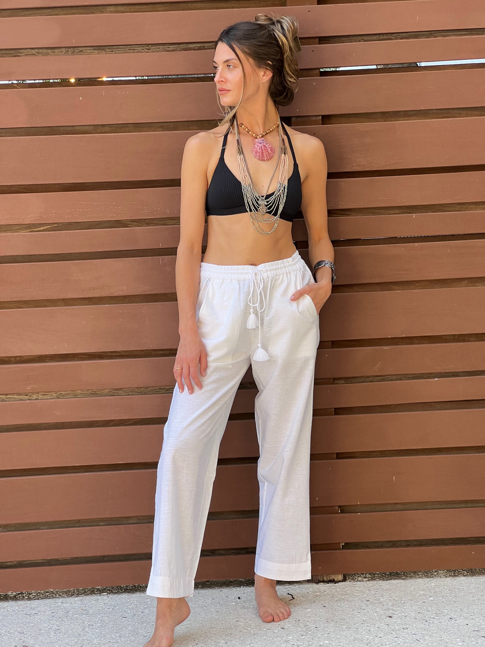 A woman standing against a wooden background wearing white wide-leg pants with a drawstring waist and front pockets, paired with a black bikini top.