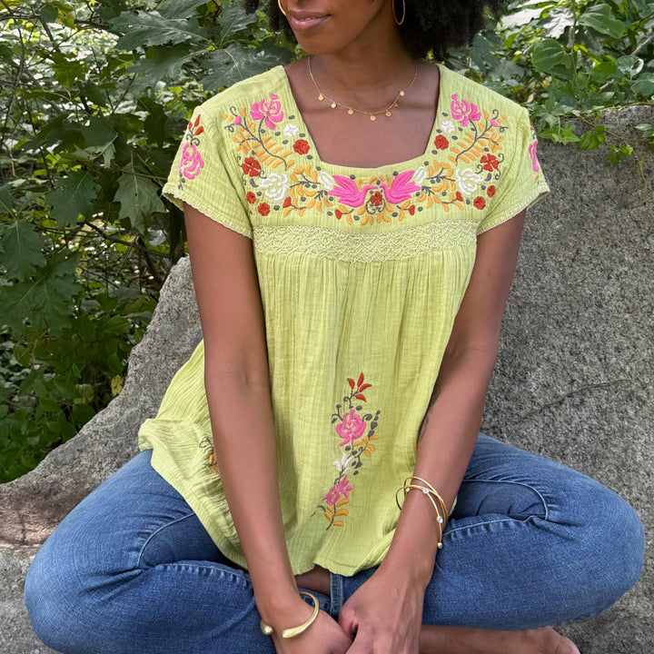 Woman sitting outdoors wearing a bright green embroidered top and blue jeans.