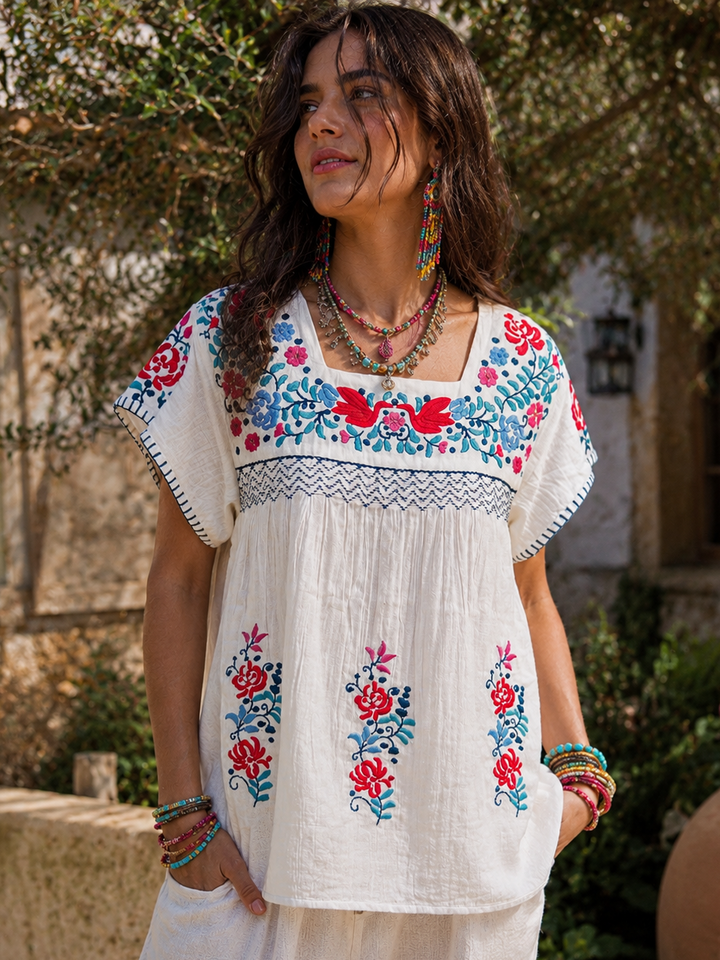 Woman wearing a white embroidered top with colorful floral patterns outdoors.